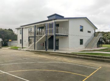 picture of school building in front of a schools court. The building is two levels painted white with stairs going up to the second level in fron.
